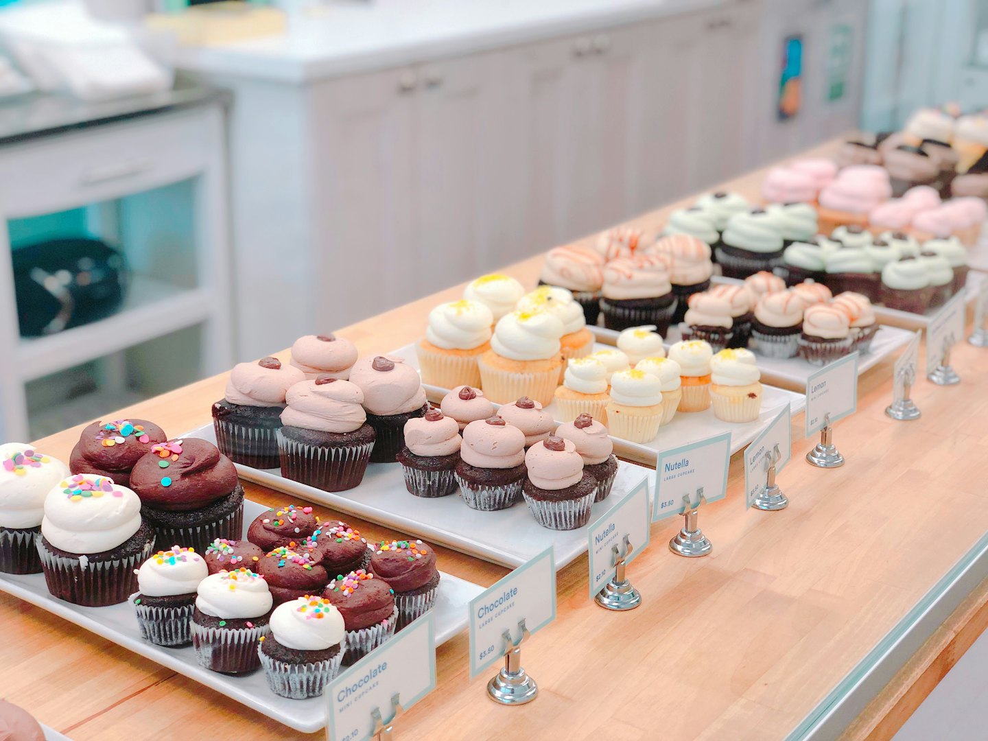 Rows of assorted cupcakes with signs on a bakery counter.